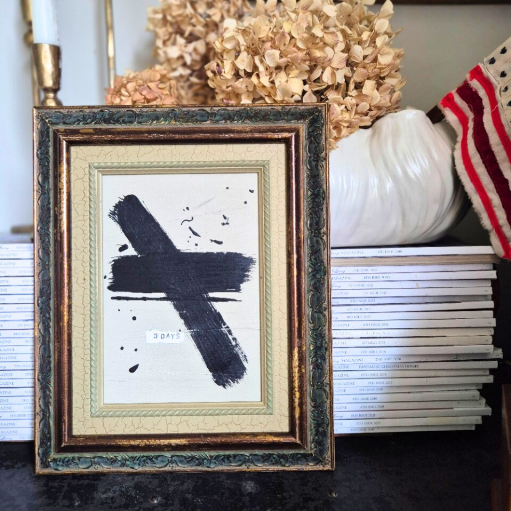 Framed abstract artwork with a black cross on a white background, placed on a surface with decorative items.