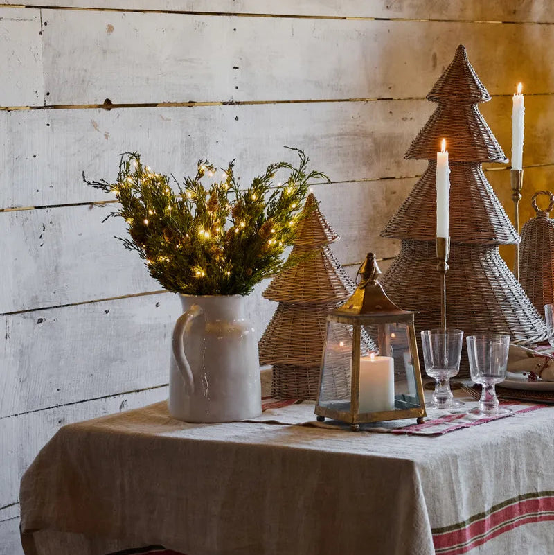 Decorative table setting with a pitcher of lights, wicker Christmas trees, and candles against a wooden wall.