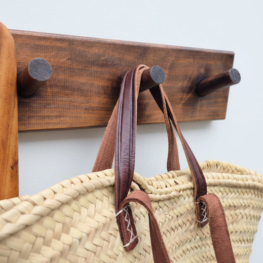Wooden wall hook with a straw bag and brown leather straps.