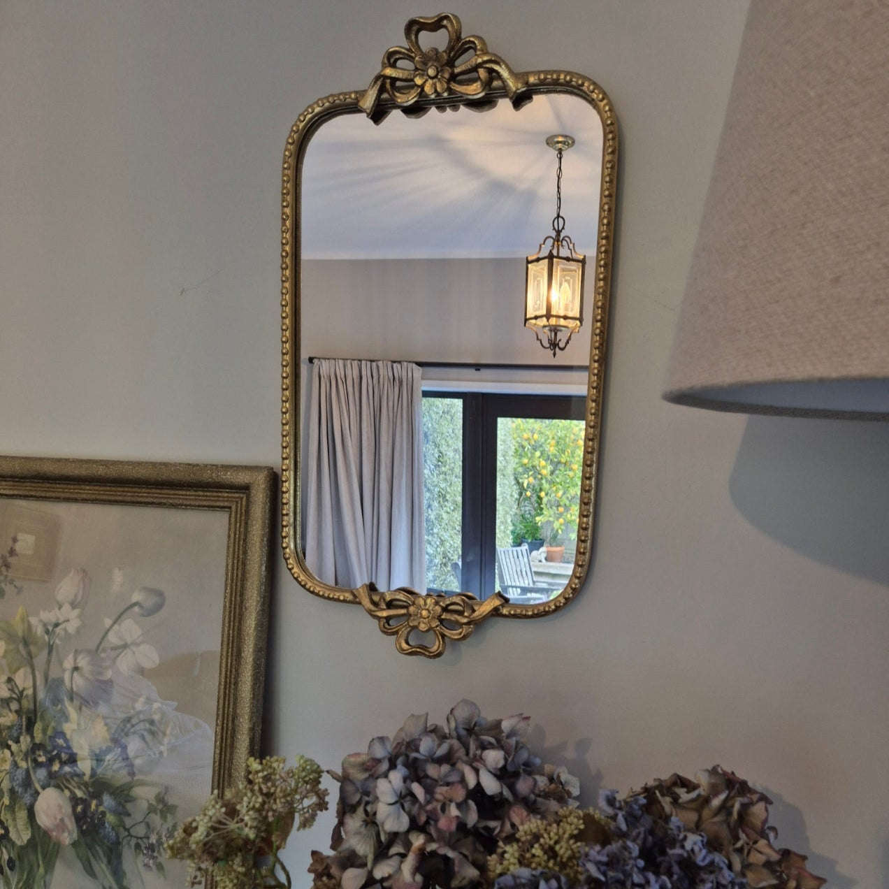 Decorative mirror with ornate frame on a wall next to a floral arrangement.
