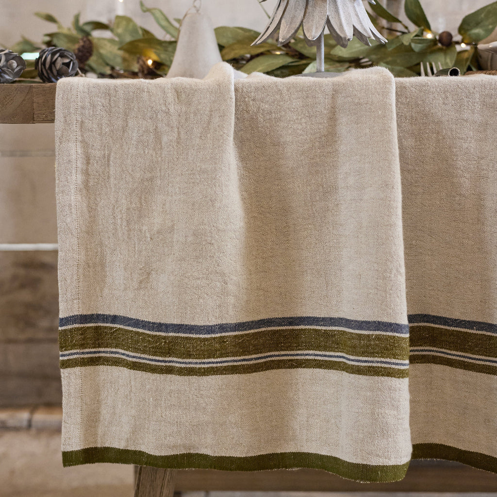 Beige Linen tablecloth with green stripes on a wooden table, set against a stone wall.