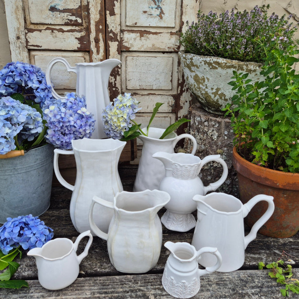 Collection of white ceramic jugs and pots with plants on a rustic wooden surface.