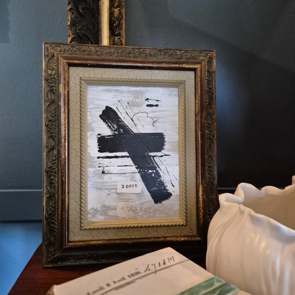 Framed artwork with a cross design on a table against a dark wall.