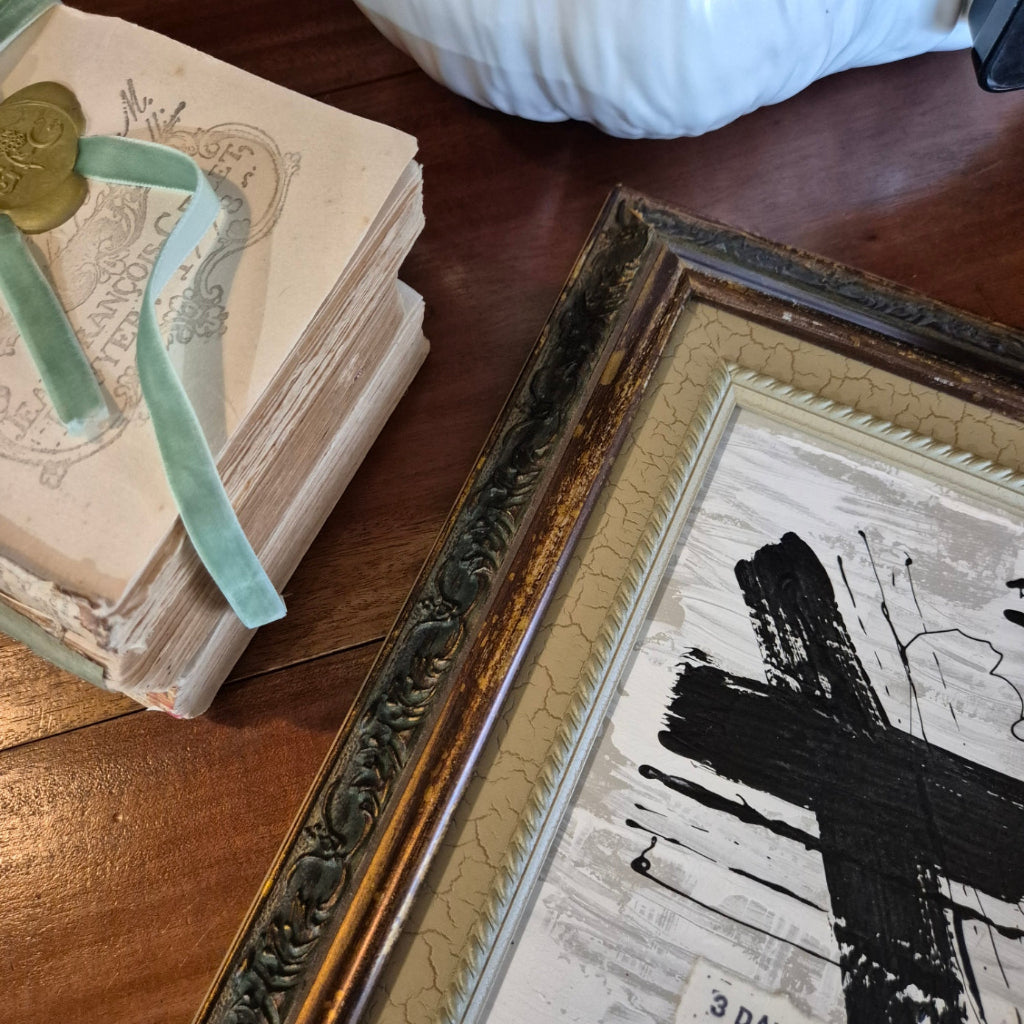 Framed artwork with a gold frame on a wooden surface next to an old book.