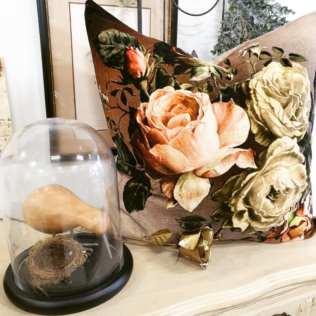 Decorative setup with a glass dome containing a gourd and nest, floral pillow in the background.