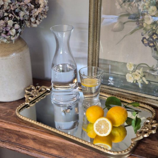 Clear carafe and glass with water on a mirrored tray with lemons and flowers in the background
