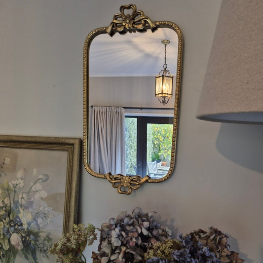 Decorative mirror with ornate frame on a wall next to a floral arrangement.