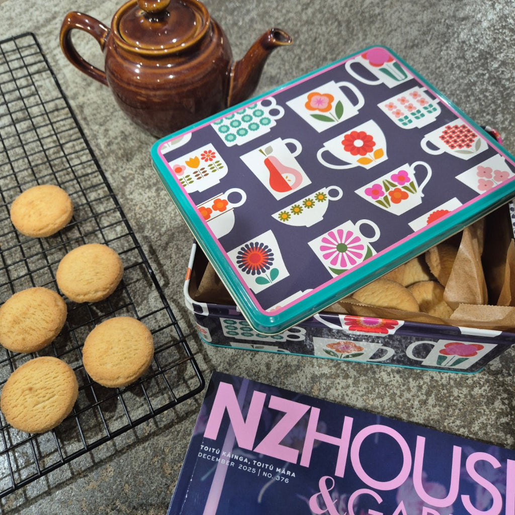 Decorative cookie tin with cookies on a cooling rack and NZ House & Garden magazine.