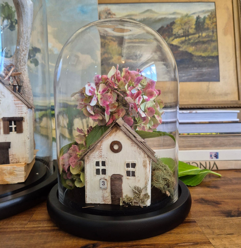 Small house model with pink flowers inside a glass dome on a wooden surface.