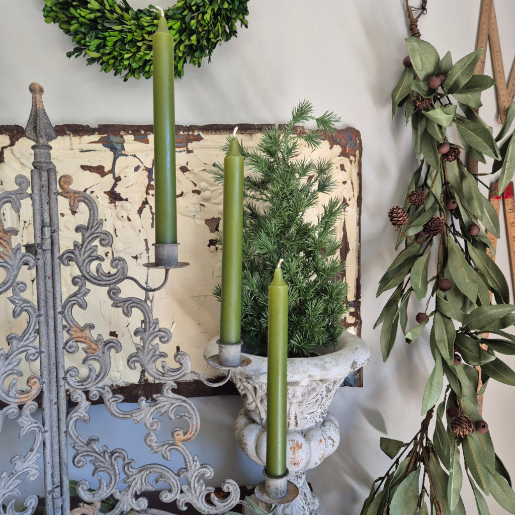 Decorative candle holder with green candles on an ornate metal stand, surrounded by greenery.