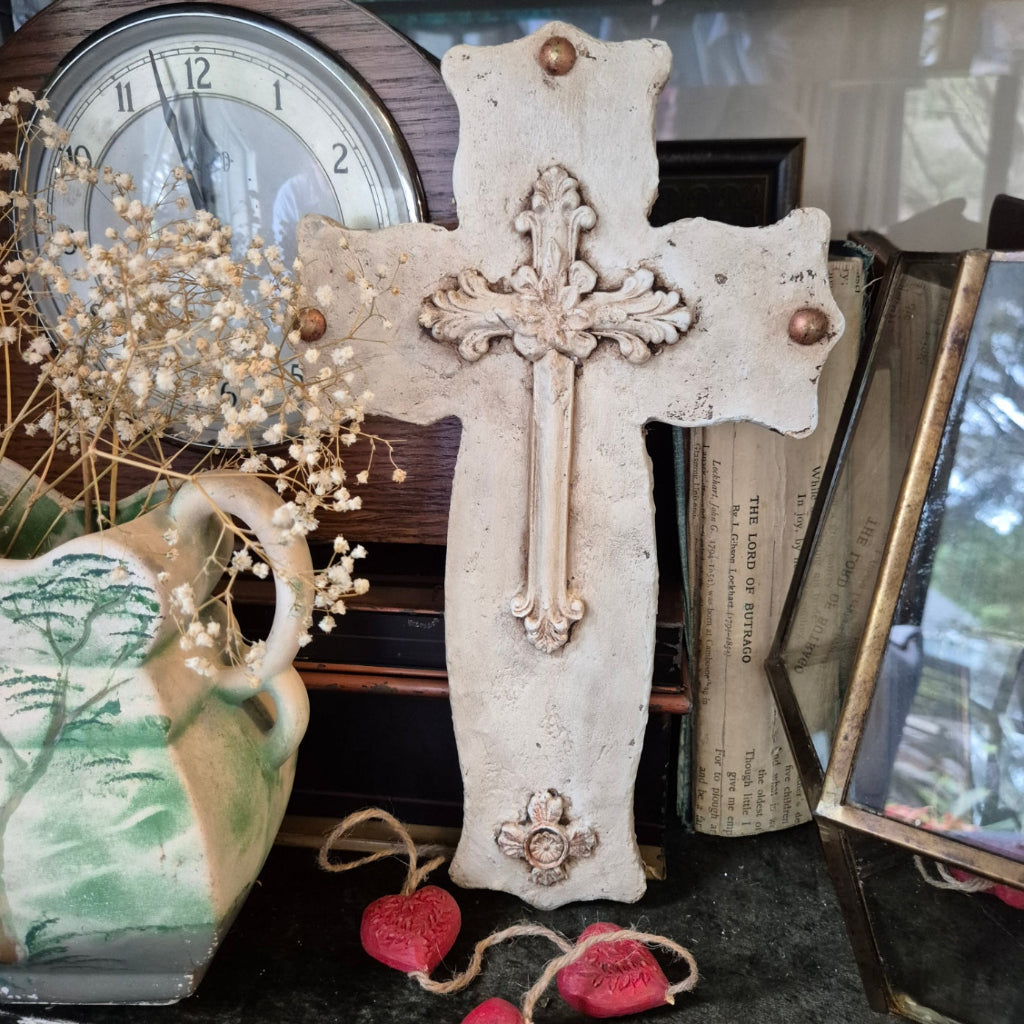 Decorative cross with ornate details on a surface with books and a clock in the background