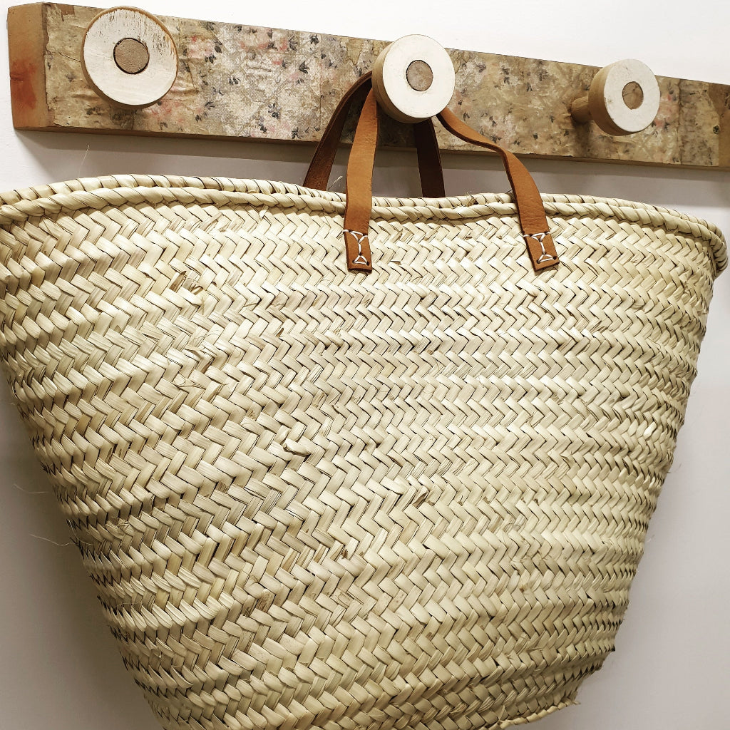 Woven basket with leather handles hanging on a wooden hook against a white wall.