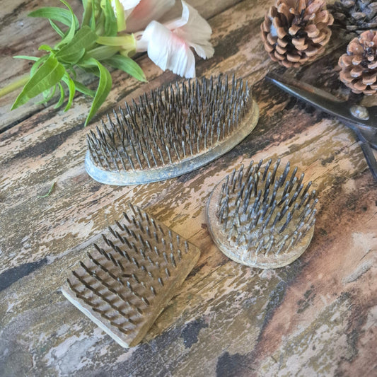 Three flower frog pins on a rustic wooden surface with pine cones and a flower.
