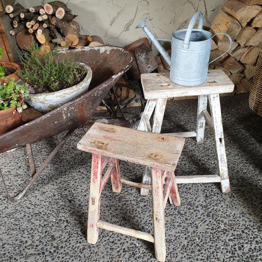 Two wooden stools and a metal watering can on a concrete floor with stacked firewood in the background.